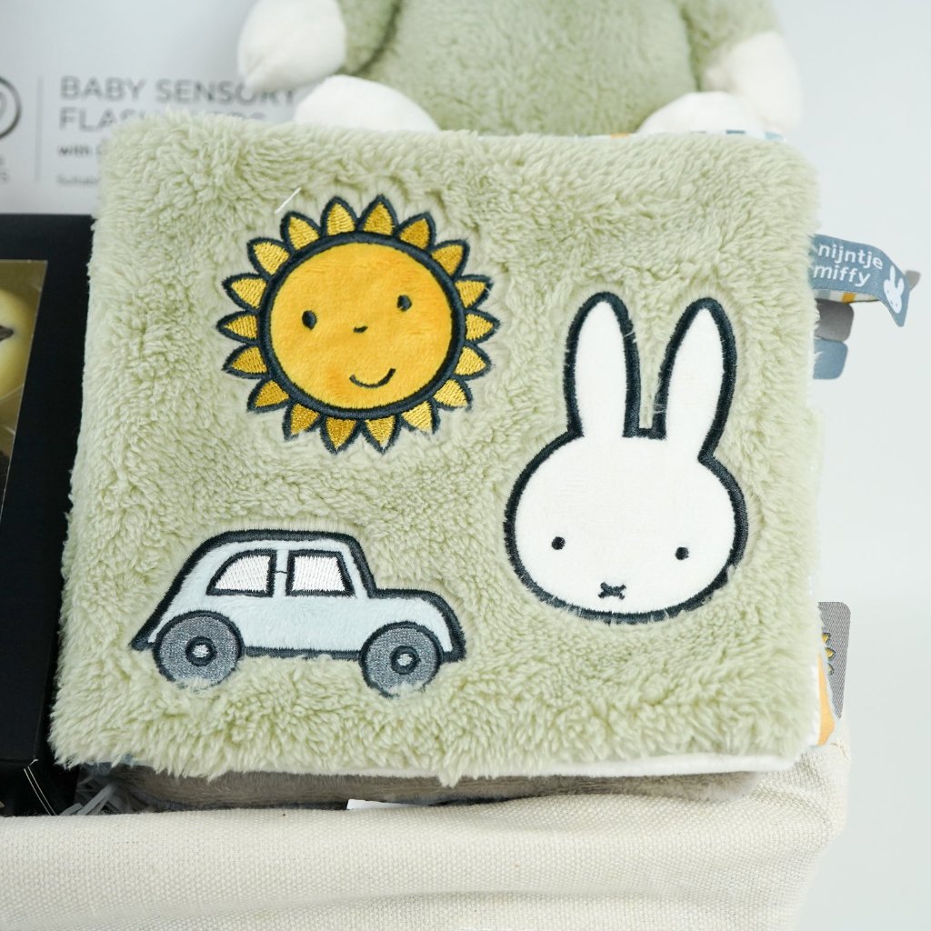 White hamper basket with baby sensory muslin, baby sensory flash cards, Sophie la Giraffe teether, Miffy soft toy, Miffy soft activity book in Green, Aromatherapy candle, chocolates
