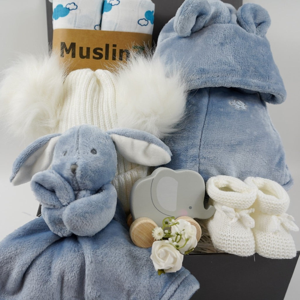 Grey magnetic box with dusty blue baby dressing gown with cute ears, dusty blue baby rabbit comforter, white baby bobble hat with 2 fluffy pom poms, grey wooden push along elephant toy, two white organic muslins with blue cloud pattern, white knit baby bootees
