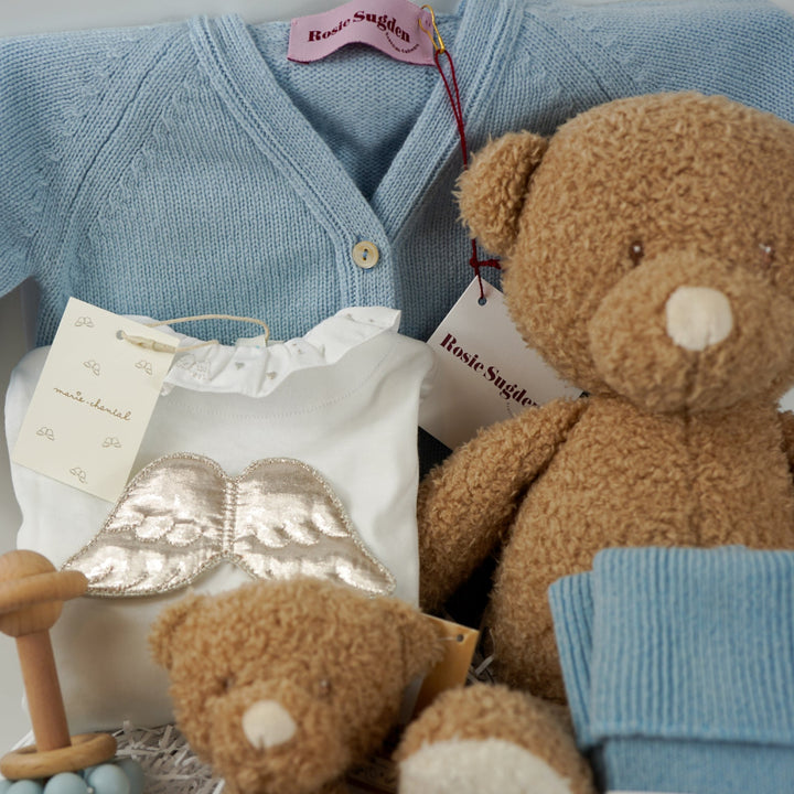 Luxury baby gift hamper box with pale blue cashmere baby cardigan, white luxury baby sleepsuit with frilled collar and silver angel wings on the back, wooden baby rattle with blue silicone teething beads, matching cashmere socks for mum in pal blue, soft teddy and matching baby comforter