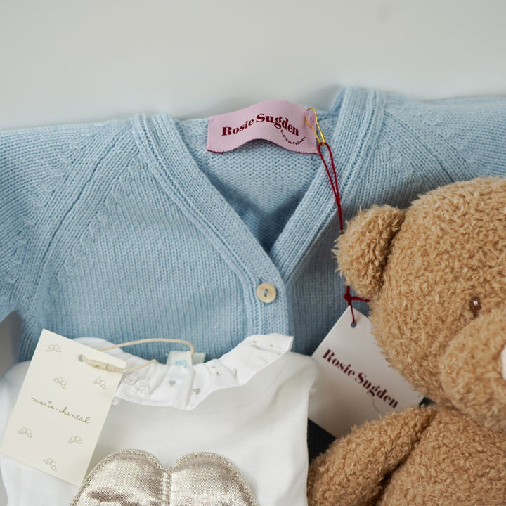 Luxury baby gift hamper box with pale blue cashmere baby cardigan, white luxury baby sleepsuit with frilled collar and silver angel wings on the back, wooden baby rattle with blue silicone teething beads, matching cashmere socks for mum in pal blue, soft teddy and matching baby comforter