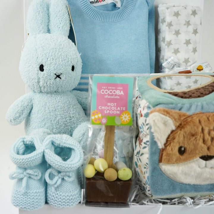 baby boy hamper with blue fine knit romper with white striped and pocket, white muslin with silver stars, blue terry miffy, Forest friends Fox soft activity book, blue knit booties, chocolate stirrer spoon with mini eggs