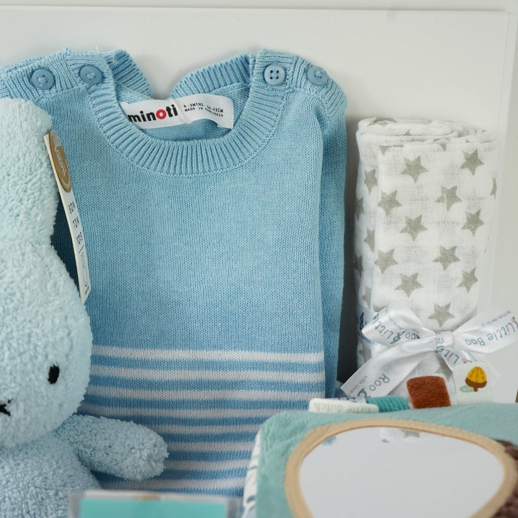 baby boy hamper with blue fine knit romper with white striped and pocket, white muslin with silver stars, blue terry miffy, Forest friends Fox soft activity book, blue knit booties, chocolate stirrer spoon with mini eggs