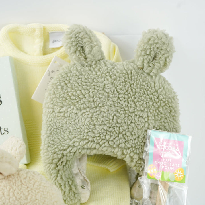 easter baby hamper, yellow knit romper with bunny design and pompom tail, soft fluffy bunny ears green hat with matching bunny ears , booties , recycled cream bunny cuddle toy, miles and moments baby cards, hot chocolate spoon with small eggs