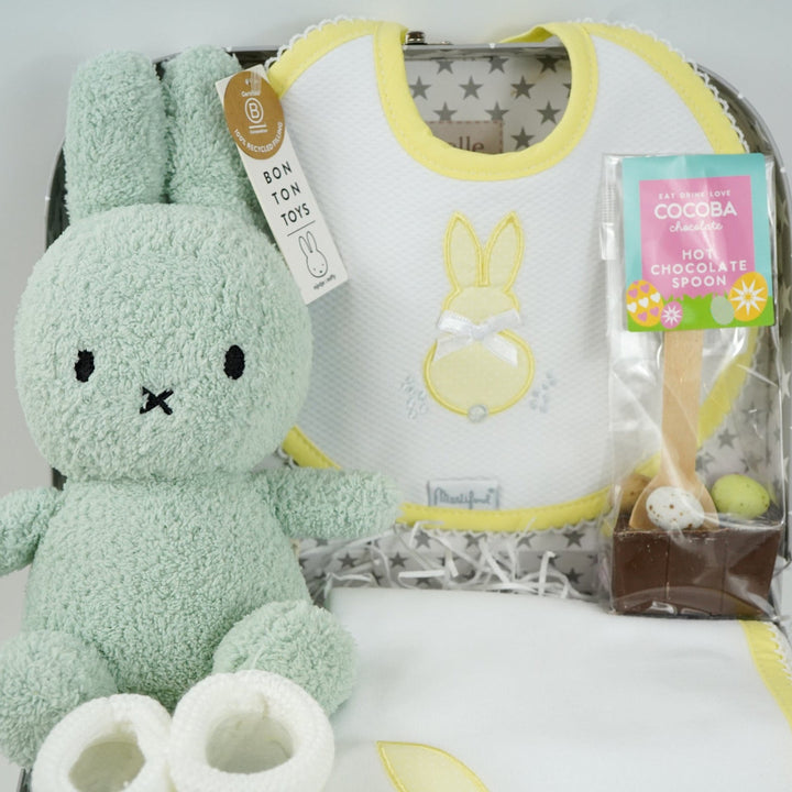 baby keepsake case with green miffy bunny, white bib with yellow scalloped edging and applique bunny with a bow and matching wrap, white baby knit booties , chocolate spoon with mini eggs