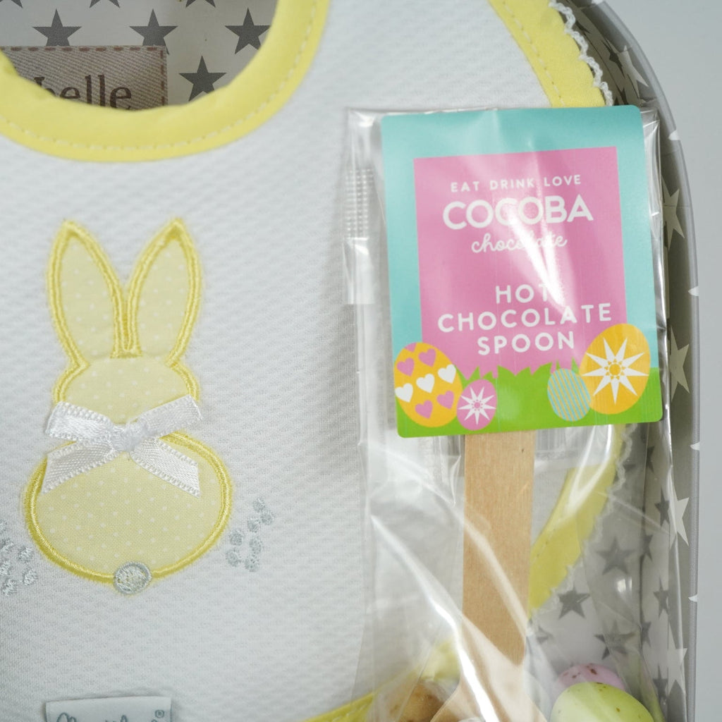 baby keepsake case with green miffy bunny, white bib with yellow scalloped edging and applique bunny with a bow and matching wrap, white baby knit booties , chocolate spoon with mini eggs
