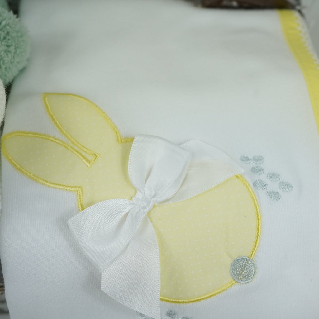 baby keepsake case with green miffy bunny, white bib with yellow scalloped edging and applique bunny with a bow and matching wrap, white baby knit booties , chocolate spoon with mini eggs