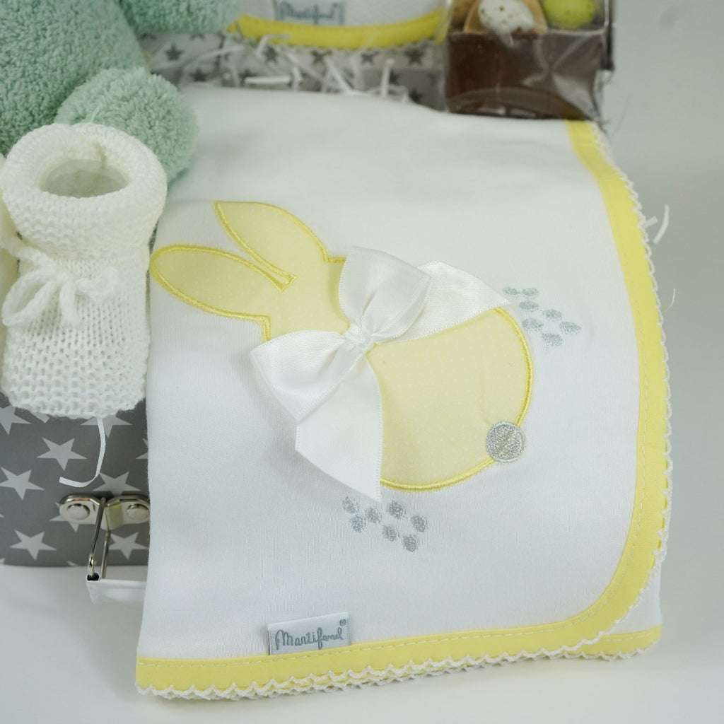 baby keepsake case with green miffy bunny, white bib with yellow scalloped edging and applique bunny with a bow and matching wrap, white baby knit booties , chocolate spoon with mini eggs