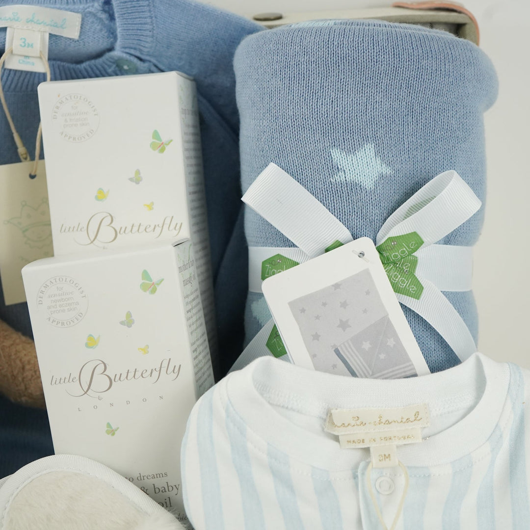 luxury baby keepsake case with luxury gifts, Steiff organic teddy with blue rompers , blue cashmere baby romper, baby toiletries organic, blue cotton blanket with star design, blue and white striped romper in organic cotton with embroidery on both sleeves, fluffy cream baby slip on slippers, wooden rattle with blue beads