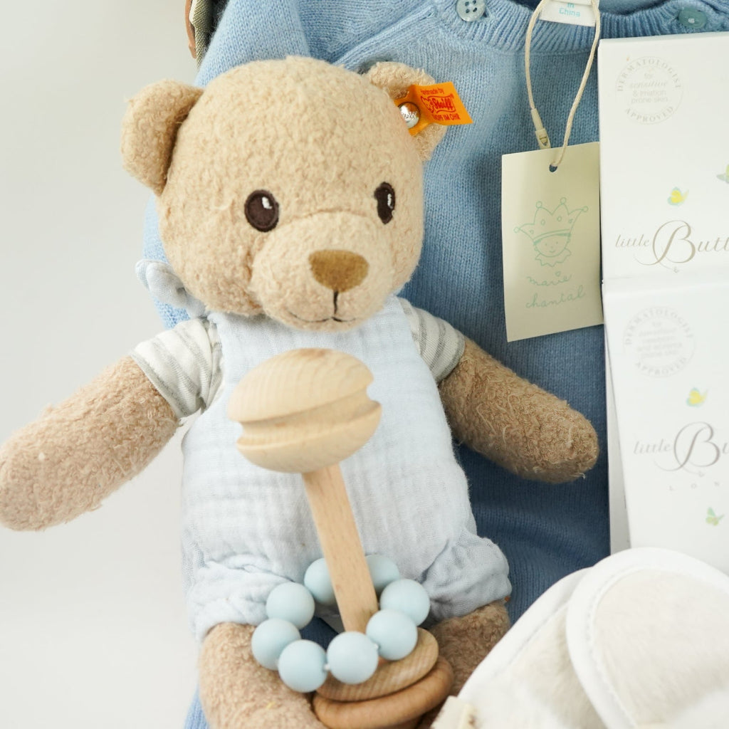 luxury baby keepsake case with luxury gifts, Steiff organic teddy with blue rompers , blue cashmere baby romper, baby toiletries organic, blue cotton blanket with star design, blue and white striped romper in organic cotton with embroidery on both sleeves, fluffy cream baby slip on slippers, wooden rattle with blue beads