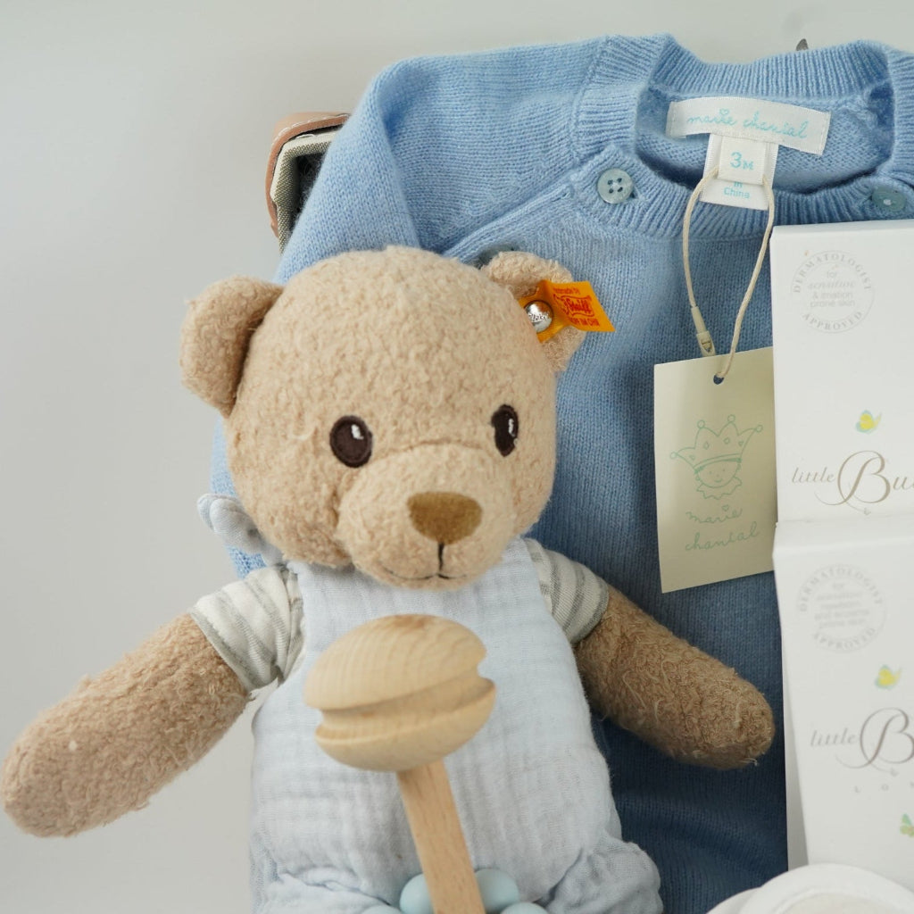 luxury baby keepsake case with luxury gifts, Steiff organic teddy with blue rompers , blue cashmere baby romper, baby toiletries organic, blue cotton blanket with star design, blue and white striped romper in organic cotton with embroidery on both sleeves, fluffy cream baby slip on slippers, wooden rattle with blue beads