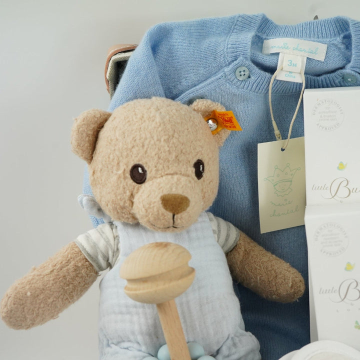 luxury baby keepsake case with luxury gifts, Steiff organic teddy with blue rompers , blue cashmere baby romper, baby toiletries organic, blue cotton blanket with star design, blue and white striped romper in organic cotton with embroidery on both sleeves, fluffy cream baby slip on slippers, wooden rattle with blue beads