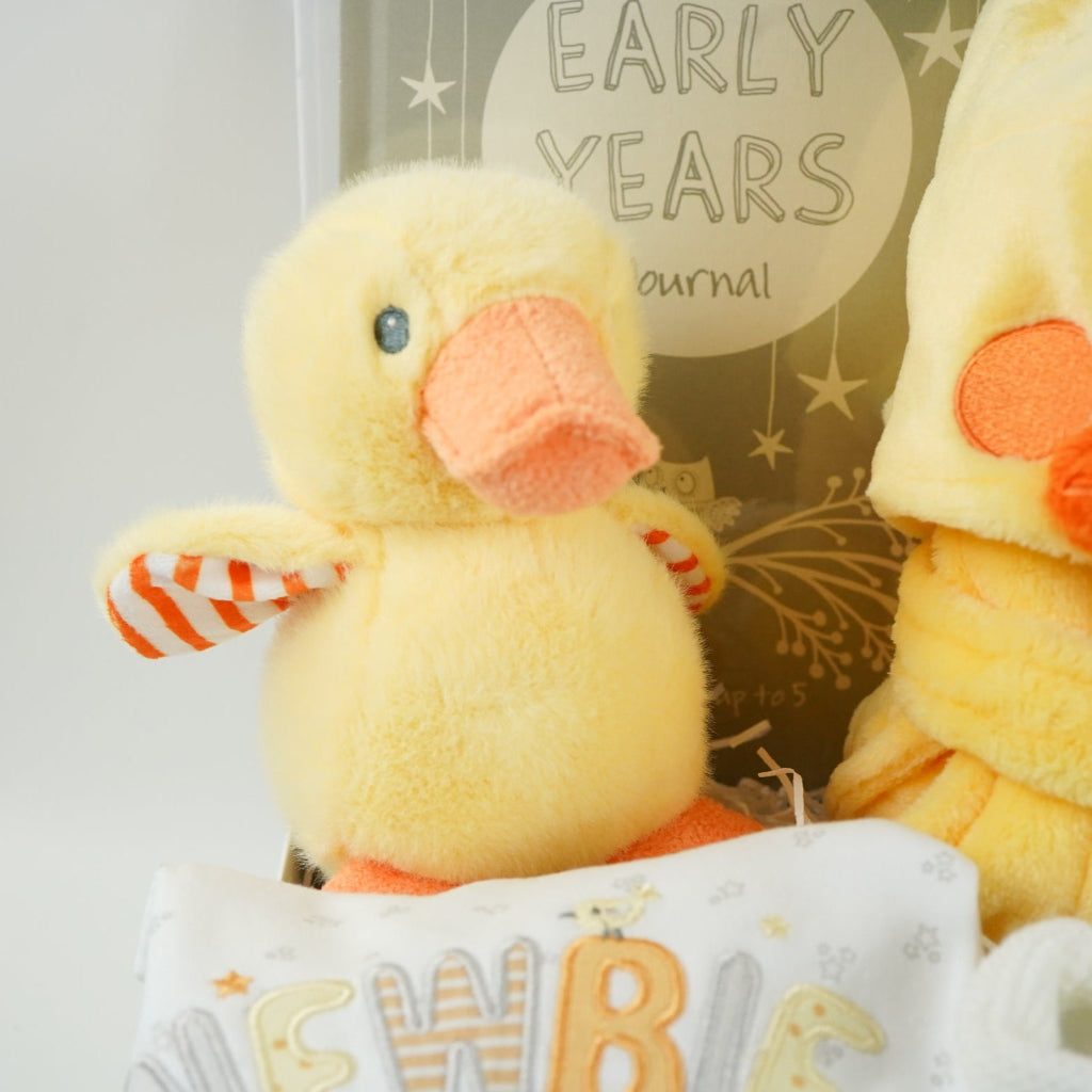 white hamper box with yellow baby dressing gown with duck face hood, soft cuddly yellow duck toy, white muslin square with yellow duck pattern., white knit baby booties, white baby sleep suit with zebra, giraffe and elephant and applique NEWBIE all in grey, yellow and orange, Early years baby journal