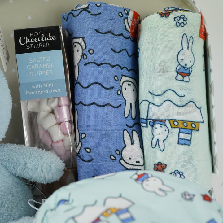 White storage suitcase with soft blue terry Miffy, pale blue knit booties with tie, salted caramel chocolate stirrer, two muslins with Miffy pattern, bucket sunhat for baby with Miffy pattern
