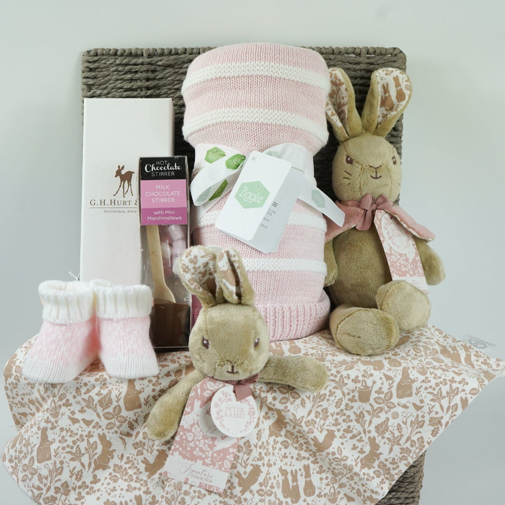 grey paper rope hamper with luxury baby brush in gift box, chocolate stirrer spoon, pink and white striped baby blanket, pink knit baby booties, peter rabbit soft toy and matching baby comforter