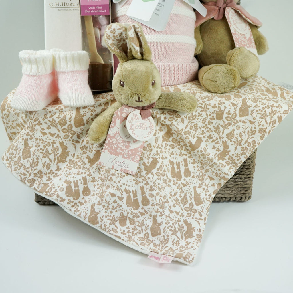 grey paper rope hamper with luxury baby brush in gift box, chocolate stirrer spoon, pink and white striped baby blanket, pink knit baby booties, peter rabbit soft toy and matching baby comforter