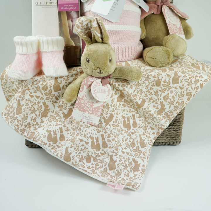 grey paper rope hamper with luxury baby brush in gift box, chocolate stirrer spoon, pink and white striped baby blanket, pink knit baby booties, peter rabbit soft toy and matching baby comforter