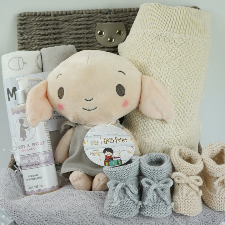 grey hamperbasket with neutral baby items including grey and cream muslin swaddles, baby toiletries, Harry Potter Dobby soft toy, grey taggie comforter , cream soft cotton baby blanket, grey booties and beige baby booties