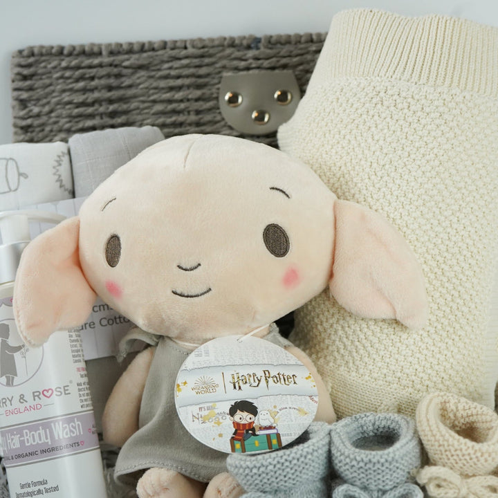 grey hamperbasket with neutral baby items including grey and cream muslin swaddles, baby toiletries, Harry Potter Dobby soft toy, grey taggie comforter , cream soft cotton baby blanket, grey booties and beige baby booties