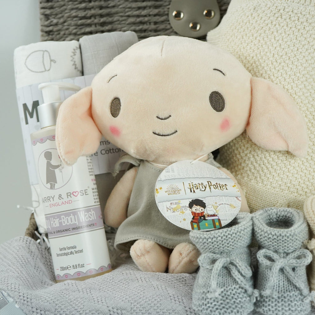 grey hamperbasket with neutral baby items including grey and cream muslin swaddles, baby toiletries, Harry Potter Dobby soft toy, grey taggie comforter , cream soft cotton baby blanket, grey booties and beige baby booties