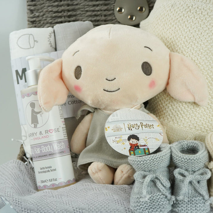 grey hamperbasket with neutral baby items including grey and cream muslin swaddles, baby toiletries, Harry Potter Dobby soft toy, grey taggie comforter , cream soft cotton baby blanket, grey booties and beige baby booties
