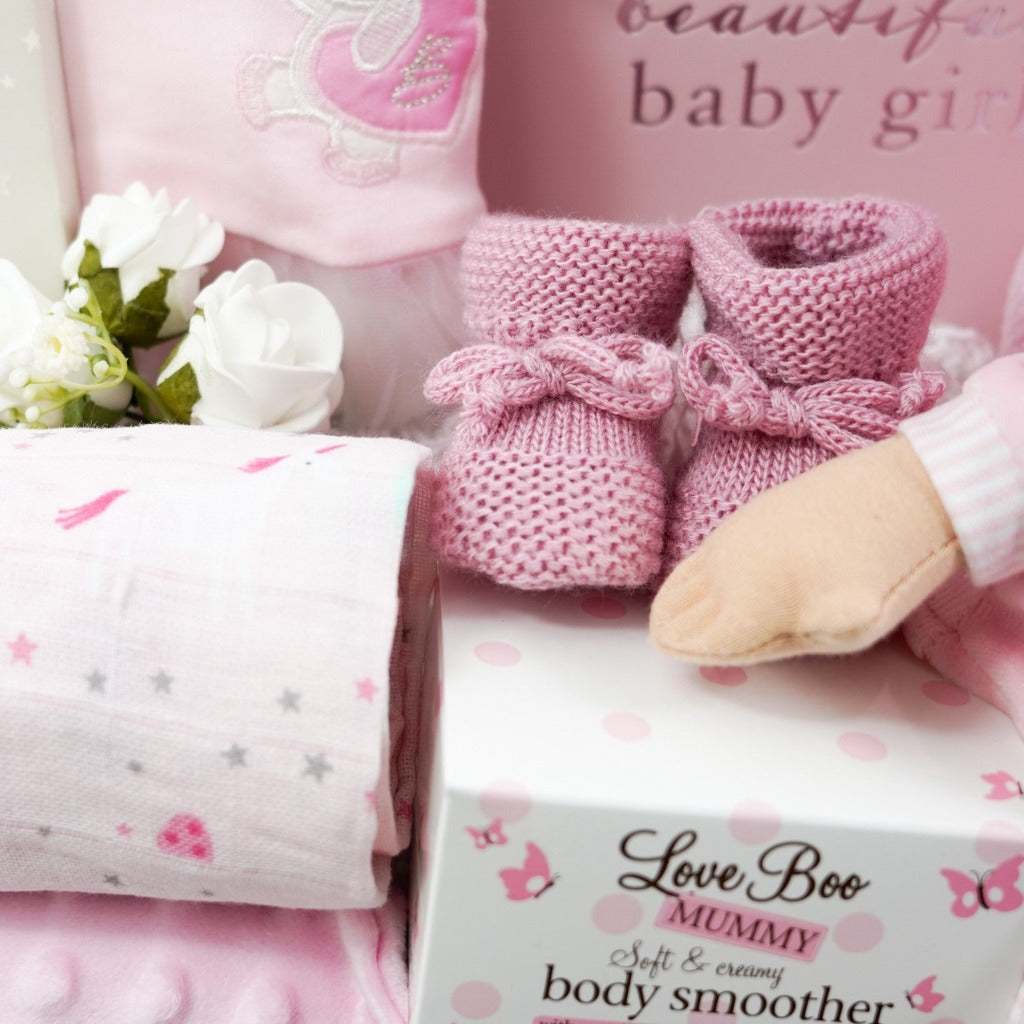 White hamper basket with Pink spanish style baby sleepsuit, baby soft doll in pink and white clothing, baby pink photo album, pink booties, baby comforter , Milestone cards , baby cellular blanket