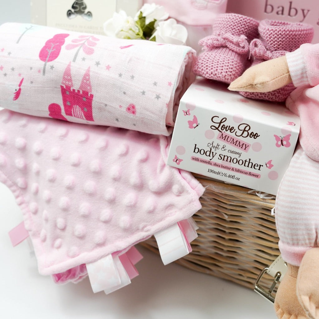 White hamper basket with Pink spanish style baby sleepsuit, baby soft doll in pink and white clothing, baby pink photo album, pink booties, baby comforter , Milestone cards , baby cellular blanket
