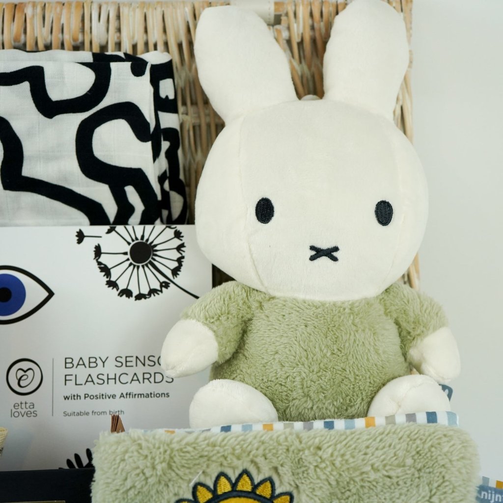 White hamper basket with baby sensory muslin, baby sensory flash cards, Sophie la Giraffe teether, Miffy soft toy, Miffy soft activity book in Green, Aromatherapy candle, chocolates