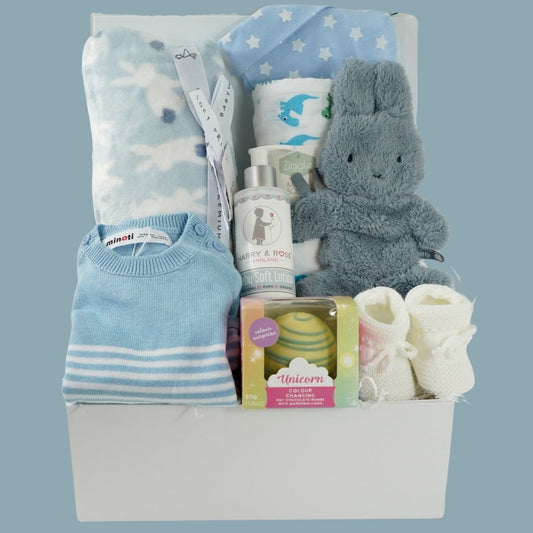 baby boy hamper box with blue and white striped fine knit baby romper, blue baby blanket with white bunnies, white muslin with blue and green dinosaurs, fluffy blue miffy comforter with dummy tag, white knit booties, organic baby soft lotion in a pump dispenser, white hot chocolate bombe