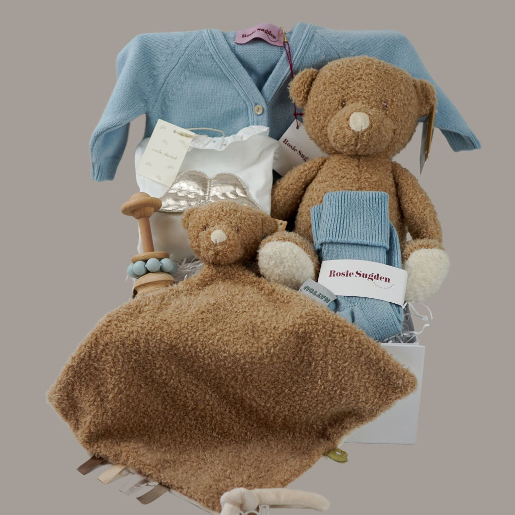 Luxury baby gift hamper box with pale blue cashmere baby cardigan, white luxury baby sleepsuit with frilled collar and silver angel wings on the back, wooden baby rattle with blue silicone teething beads, matching cashmere socks for mum in pal blue, soft teddy and matching baby comforter