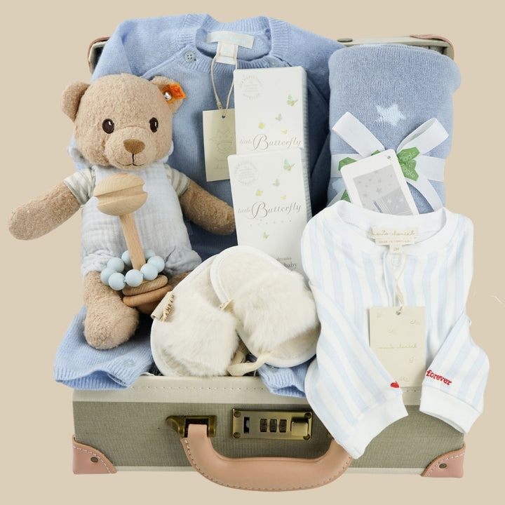 luxury baby keepsake case with luxury gifts, Steiff organic teddy with blue rompers , blue cashmere baby romper, baby toiletries organic, blue cotton blanket with star design, blue and white striped romper in organic cotton with embroidery on both sleeves, fluffy cream baby slip on slippers, wooden rattle with blue beads
