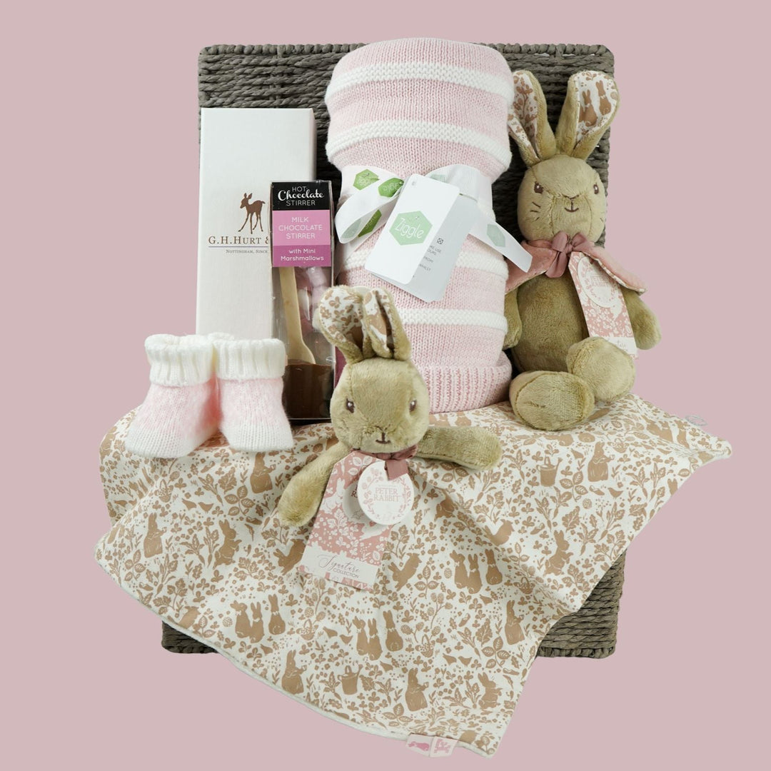 grey paper rope hamper with luxury baby brush in gift box, chocolate stirrer spoon, pink and white striped baby blanket, pink knit baby booties, peter rabbit soft toy and matching baby comforter