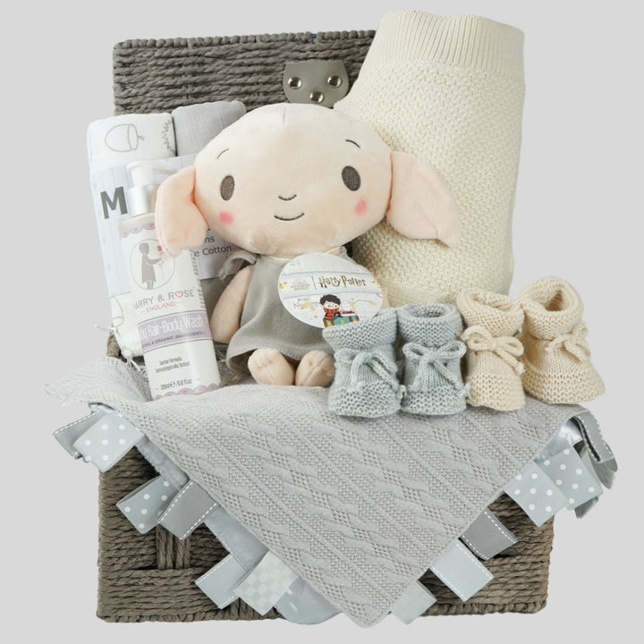 grey hamperbasket with neutral baby items including grey and cream muslin swaddles, baby toiletries, Harry Potter Dobby soft toy, grey taggie comforter , cream soft cotton baby blanket, grey booties and beige baby booties