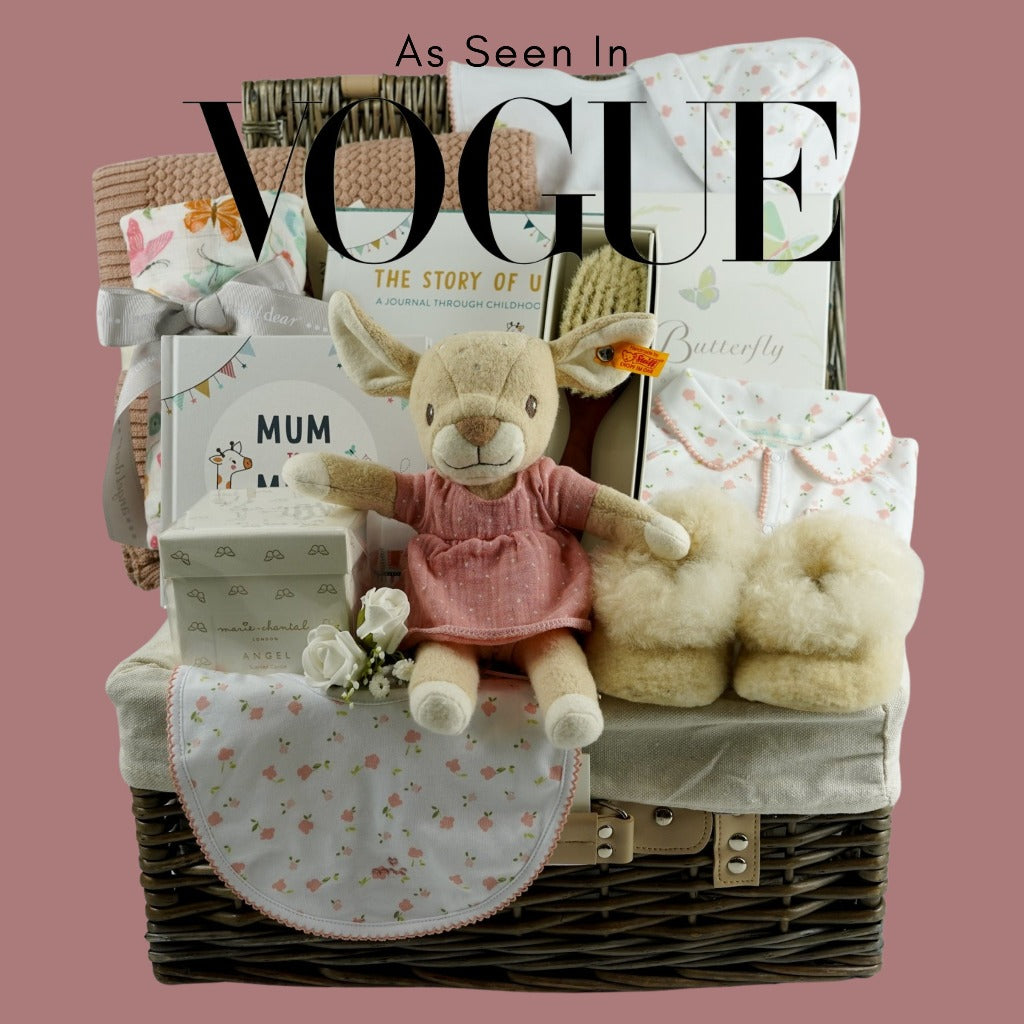 Hamper basket with luxury baby gifts as seen in Vogue, baby blanket, Little Butterfly london baby toiletries, Marie Chantal white candle in a box with gold angel wings, Marie chantal white baby clothing set with small pink flowers , Deer soft toy by Steiff in a pink dress, Marie Chantal baby luxury hairbrush, Luxury baby champagne coloured alpaca slippers