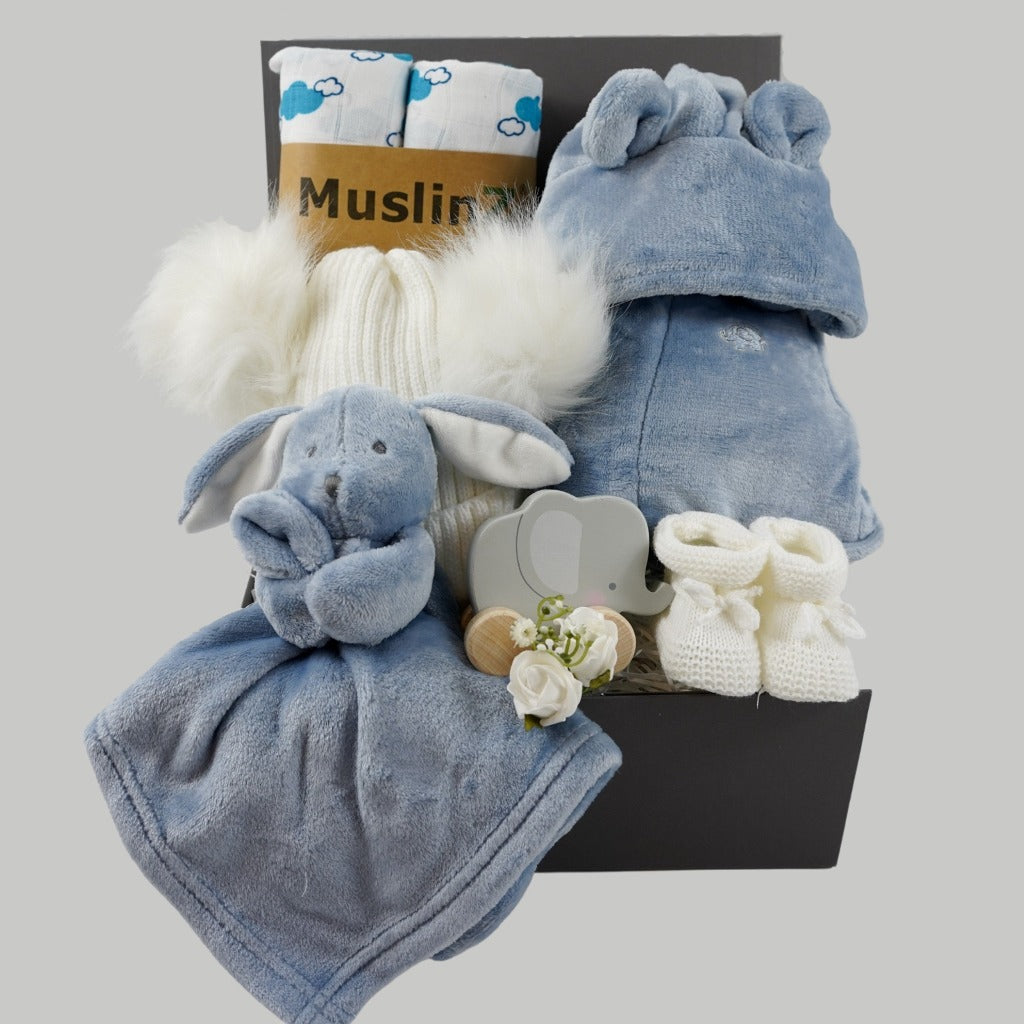 Grey magnetic box with dusty blue baby dressing gown with cute ears, dusty blue baby rabbit comforter, white baby bobble hat with 2 fluffy pom poms, grey wooden push along elephant toy, two white organic muslins with blue cloud pattern, white knit baby bootees