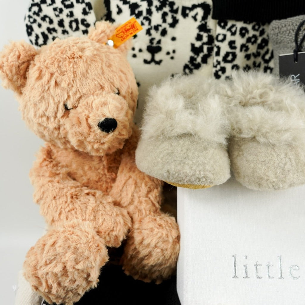 baby hamper basket with honey coloured steiff teddy, baby alpaca slippers in champagne, baby knitted jumper in organic cream cotton with black leopard design, black baby cotton knitted leggings, black cotton knit baby hat with ears, baby photo album in white linen, baby stacking ring toy with rabbit head, grey cotton baby cellular blanket