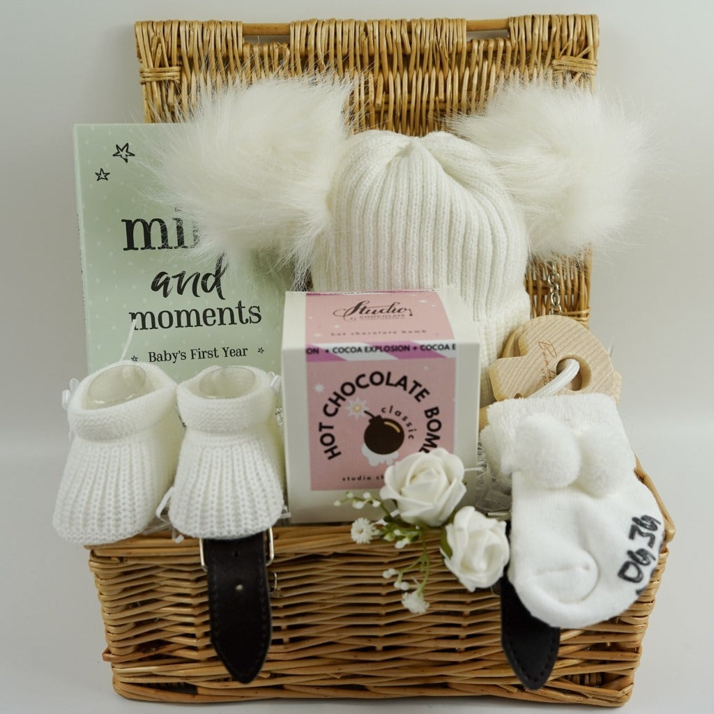 Small hamper basket with baby milestone cards in a box, white knit hat with 2 fluffy pom poms, baby booties in white, white baby socks with a cute message on the soles, wooden teething keys , Hot chocolate bomb