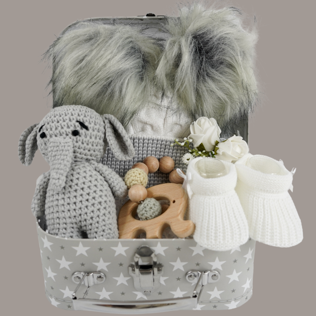 Grey gift suitcase with white stars, cream baby hat with grey trim and grey fluffy double pom poms , grey amigurumi elephant and wooden elephant teether with wooden and crocheted beads , white baby booties