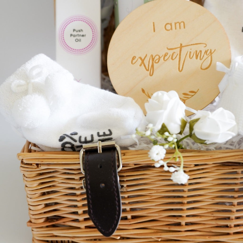 small wicker hamper, perineal oil, wooden pregnancy milestone discs in a cream bag, white with grey elephants baby muslin, white with silver stars baby knot hat, baby socks with cute writing , baby booties