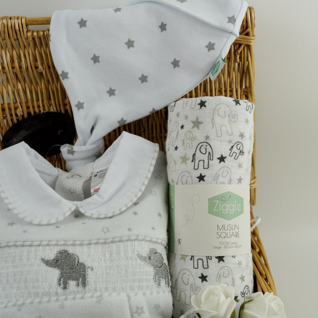 Small wicker basket, white velour baby sleepsuit with stars and grey elephants with a smocked front, white muslin with grey elephants, white cotton baby knot hat with silver stars
