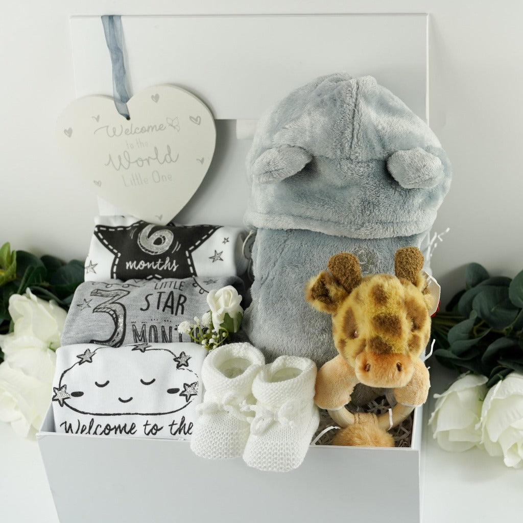 white baby hamper in a ribbon tabbed magnetic box, includes a soft grey baby dressing gown with cute ears baby three milestone vests in white, grey and black , marking baby's arrib=vasl, 3 months and 6 months, white knit booties, giraffe soft rattle and white nursery plaque with welcome to the world in silver writing