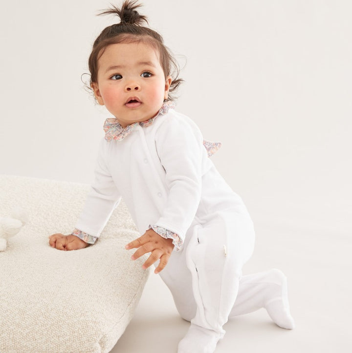 White velour baby sleepsuit with liberty print ruffle collar and libertiy print wings and trims around the cuffs, the sleepsuit is a soft velour