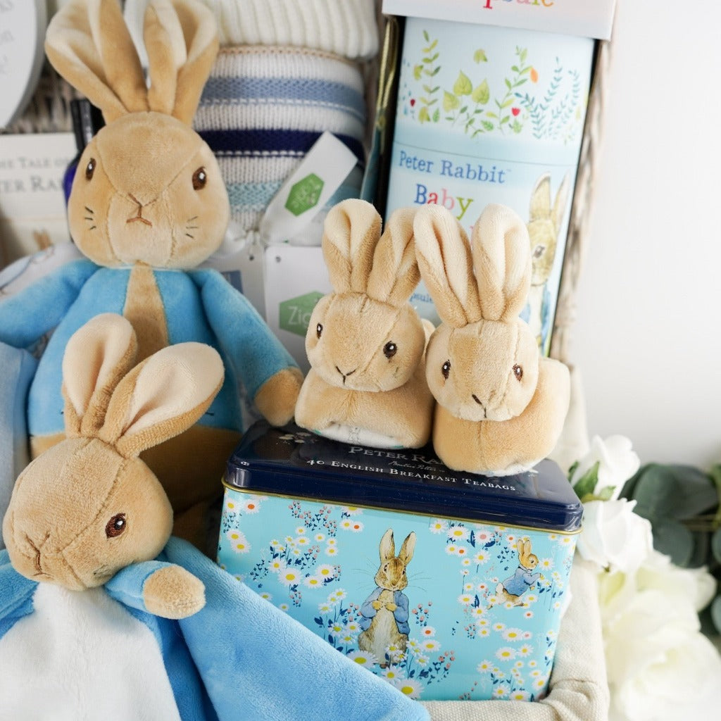 Luxury baby hamper with Peter rabbit itmes including Peter Rabbit soft toy, Peter rabbit baby comforter in blue and white, Soft brown Peter Rabbit baby slippers, Peter Rabbit Time Capsule, Peter Rabbit Book, Peter Rabbit baby clothing set including a pale blue jacket with Peter Rabbit embroidered, Luxury chocolate bar, collectors Peter Rabbit tin with breakfast tea, Soft blue and beige stripe heavy knit baby blanket, Neals yard baby toiletries
