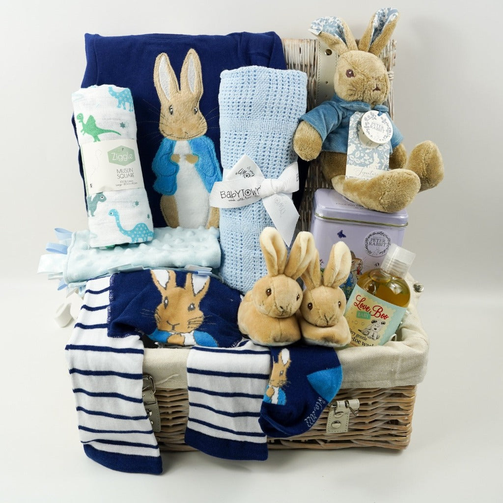 Peter rabbit themed baby basket with navy clothing set with a top with PPeter Rabbit applique design, stiped leggings with Peter Rabbit on the bottom, matching softs, Peter Rabbit soft slippers, Muslin in white with green and blue dinosaurs, pale blue cotton cellular blanket, soft Peter Rabbit toy, Tin of tea with Peter Rabbit design, Natural baby toiletries