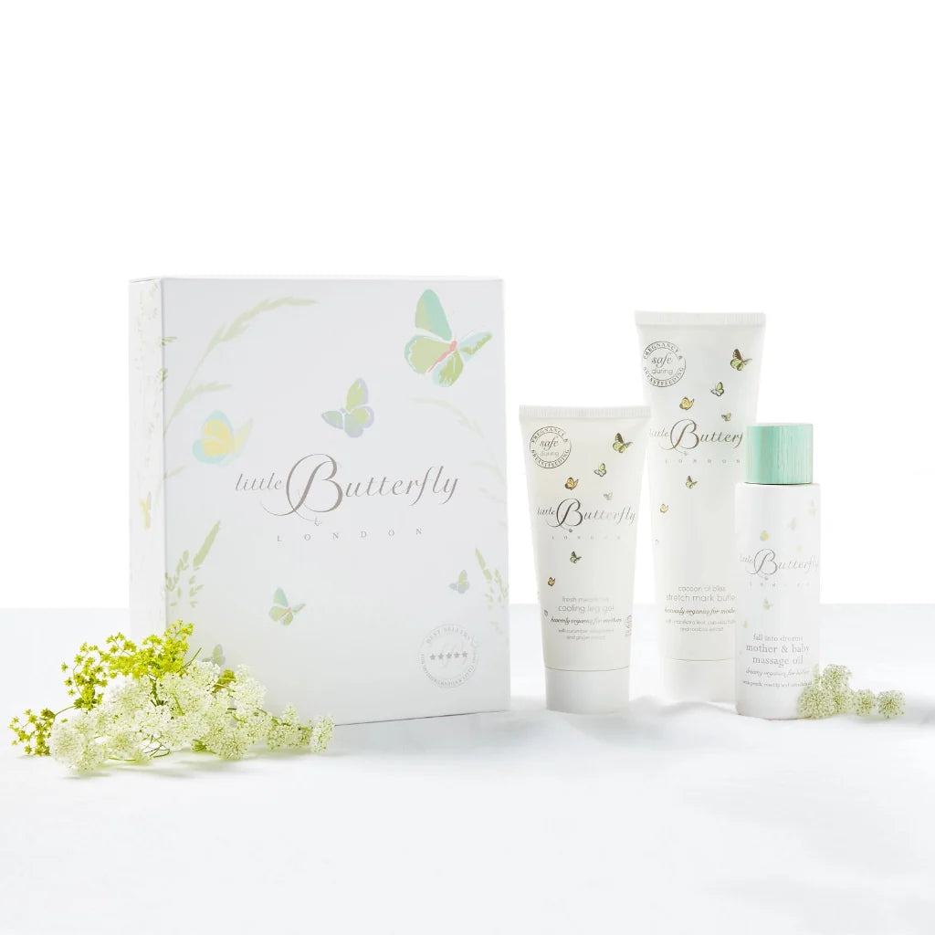 A pregnancy skincare gift set from Little Butterfly London featuring a white 'little Butterfly' box and three matching white tubes and bottles with delicate butterfly illustrations, green accents, and small white flowers placed around them 