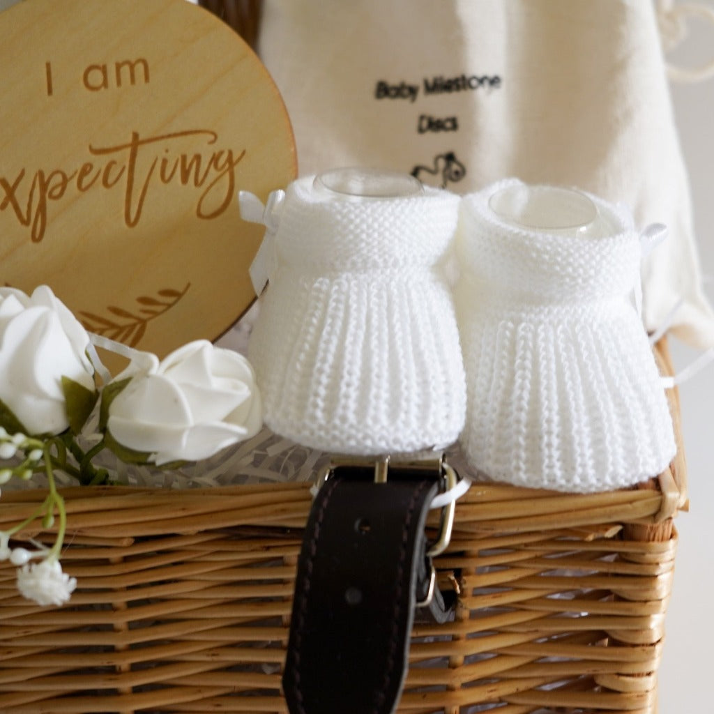 small wicker hamper, perineal oil, wooden pregnancy milestone discs in a cream bag, white with grey elephants baby muslin, white with silver stars baby knot hat, baby socks with cute writing , baby booties 