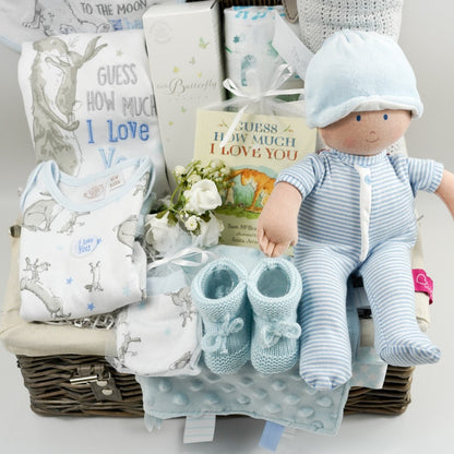 Wicker hamper with white baby set with Guess How Much I Love you Rabbit, baby rag doll in blue and white romper, grey cellular blanket, muslin , baby booties in pale blue with tie, organic baby toiletries
