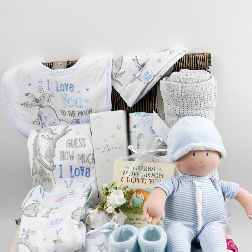 Wicker hamper with white baby set with Guess How Much I Love you Rabbit, baby rag doll in blue and white romper, grey cellular blanket, muslin , baby booties in pale blue with tie, organic baby toiletries