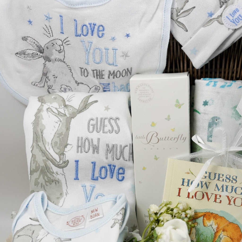 Natural hamper basket with baby 'Guess how much I love you clothing set, baby Butterfly London toiletries, muslin with green and blue dinosaurs, Grey cotton cellular blanket, baby taggie blanket, blue knit booties, Steiff Hoppie bunny in blue