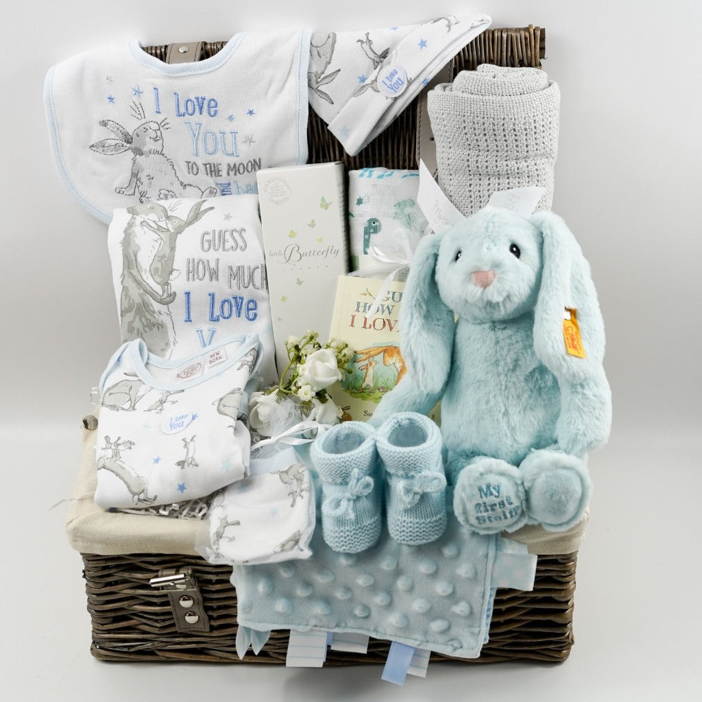 Natural hamper basket with baby 'Guess how much I love you clothing set, baby Butterfly London toiletries, muslin with green and blue dinosaurs, Grey cotton cellular blanket, baby taggie blanket, blue knit booties, Steiff  Hoppie bunny in blue