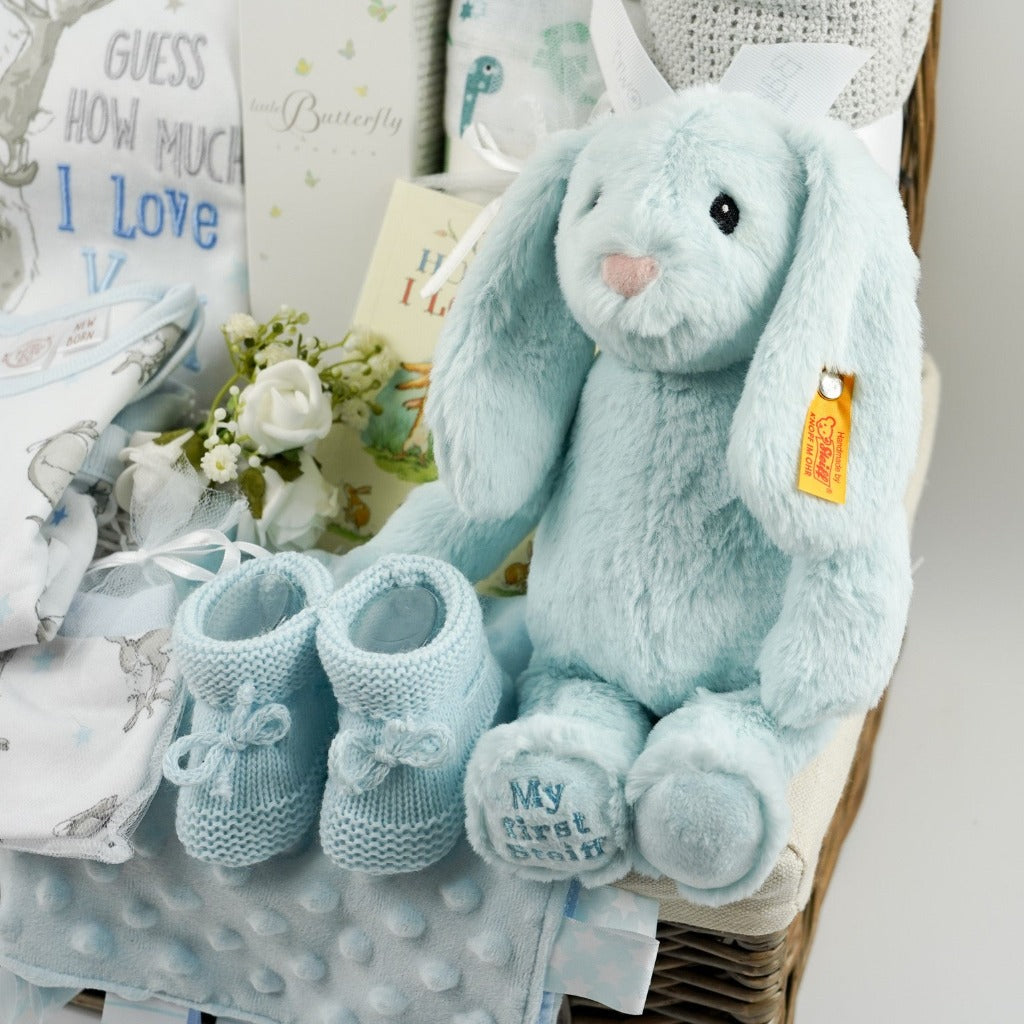 Natural hamper basket with baby 'Guess how much I love you clothing set, baby Butterfly London toiletries, muslin with green and blue dinosaurs, Grey cotton cellular blanket, baby taggie blanket, blue knit booties, Steiff Hoppie bunny in blue